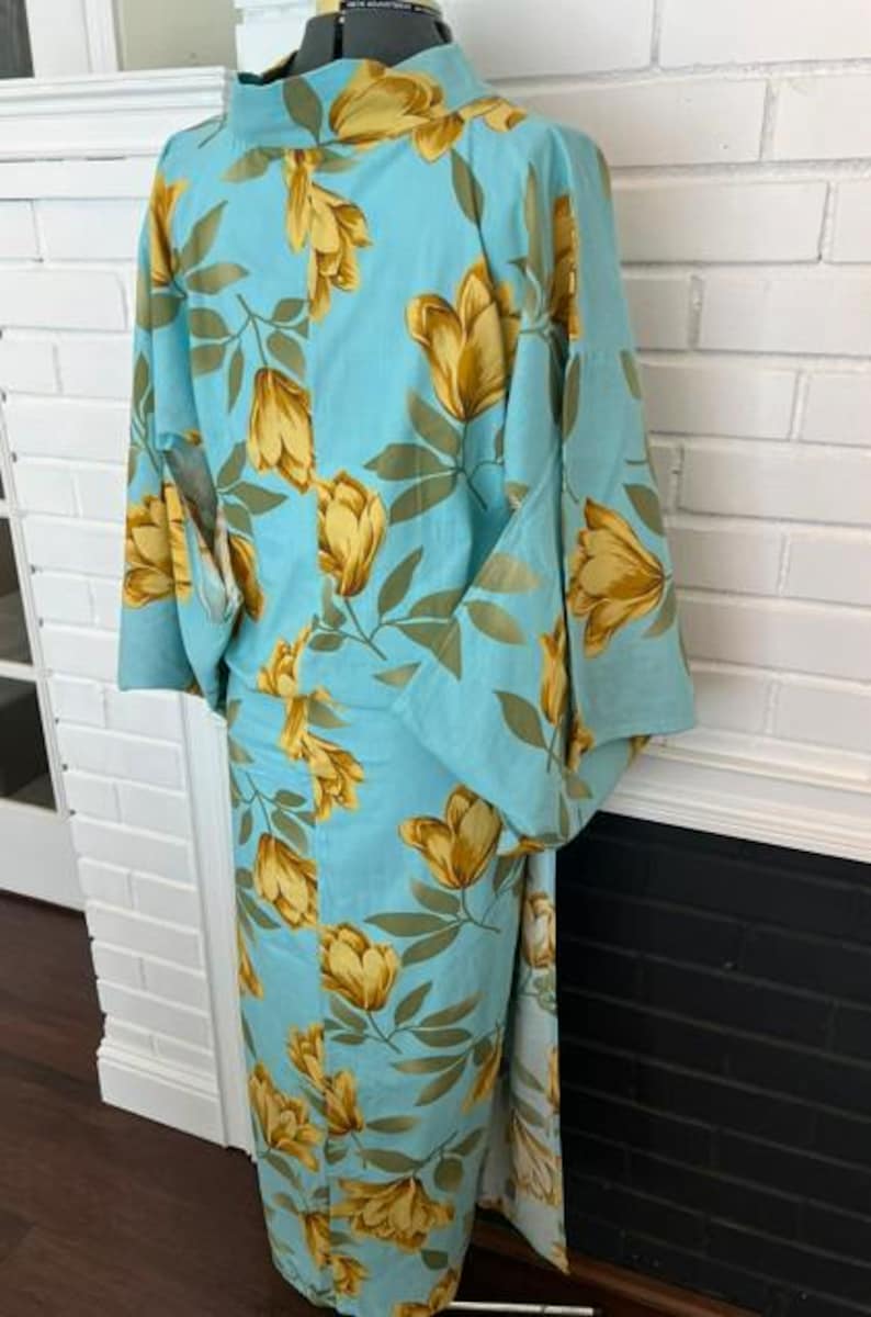 May include: A light blue kimono featuring a repeating pattern of golden yellow tulip-like flowers and muted green leaves. The garment has wide sleeves, a high collar, and a straight-cut silhouette that extends to the floor, displayed on a mannequin.