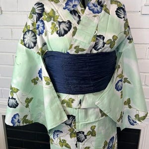 Op de afbeelding: Een licht muntgroene yukata met een herhalend patroon van blauwe en zwarte ochtendgloriebloemen met groene bladeren. Het kledingstuk is gestyled met een brede, donker marineblauwe, getextureerde obi-ceintuur rond de taille voor contrast.