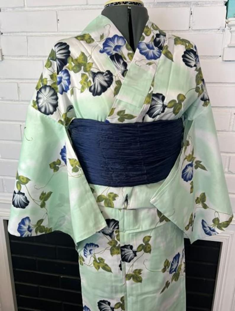 Op de afbeelding: Een licht muntgroene yukata met een herhalend patroon van blauwe en zwarte ochtendgloriebloemen met groene bladeren. Het kledingstuk is gestyled met een brede, donker marineblauwe, getextureerde obi-ceintuur rond de taille voor contrast.
