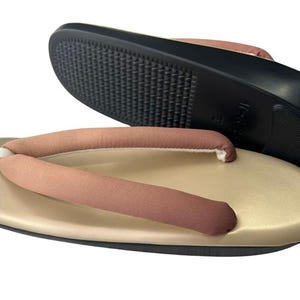 May include: Tan and brown Japanese geta sandals. The sandals have a tan base with a brown thong and a black sole. The sole has a textured pattern for grip. The sandals are designed for traditional Japanese wear.