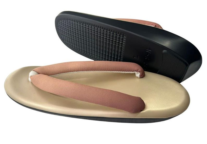 May include: Tan and brown Japanese geta sandals. The sandals have a tan base with a brown thong and a black sole. The sole has a textured pattern for grip. The sandals are designed for traditional Japanese wear.