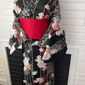 May include: A black kimono featuring a floral pattern with white and pink blossoms, red accents, and vertical geometric lines. The garment is cinched at the waist with a wide, solid red obi sash, showcasing long, wide sleeves and a wrap-style front.