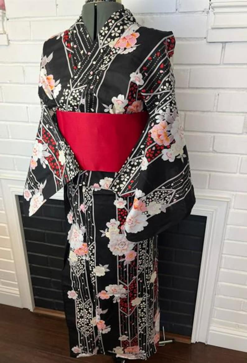 May include: A black kimono featuring a floral pattern with white and pink blossoms, red accents, and vertical geometric lines. The garment is cinched at the waist with a wide, solid red obi sash, showcasing long, wide sleeves and a wrap-style front.