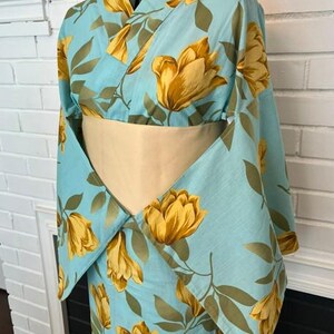 May include: A light blue kimono featuring a repeating pattern of large yellow flowers and olive green leaves. The garment has wide sleeves and is cinched at the waist with a solid beige obi sash, displayed on a mannequin torso.