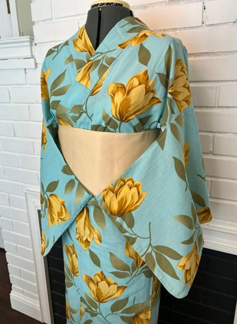 May include: A light blue kimono featuring a repeating pattern of large yellow flowers and olive green leaves. The garment has wide sleeves and is cinched at the waist with a solid beige obi sash, displayed on a mannequin torso.