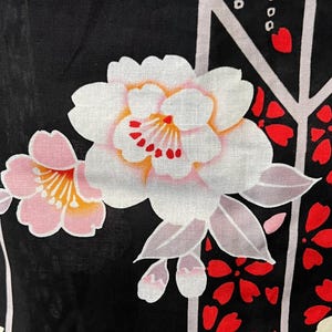 May include: A black fabric featuring a floral pattern with large white and pink blossoms, yellow centers, and red accents. The design includes light gray vertical lines and scattered red petal shapes, creating a contrast against the dark background.