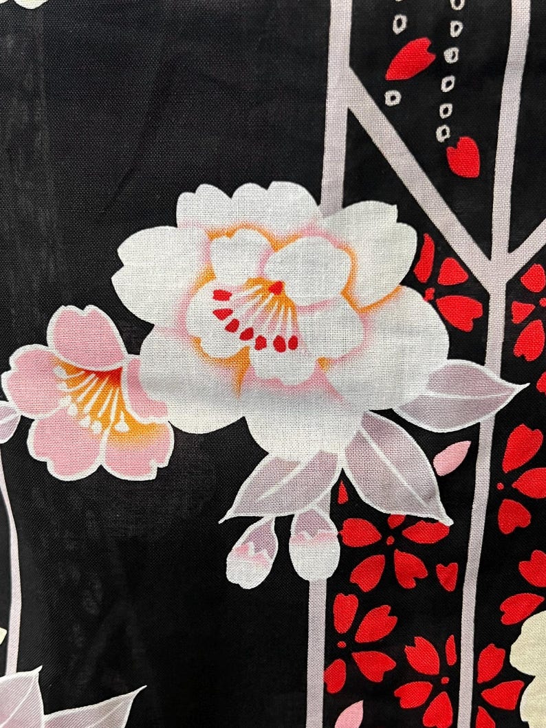 May include: A black fabric featuring a floral pattern with large white and pink blossoms, yellow centers, and red accents. The design includes light gray vertical lines and scattered red petal shapes, creating a contrast against the dark background.