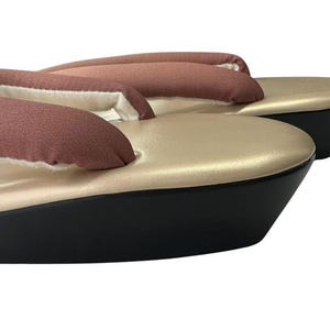 May include: A pair of traditional Japanese geta sandals. The sandals feature a gold-colored platform with a black base. The thong straps are a muted reddish-brown color. The sandals are elevated and appear to be unworn.