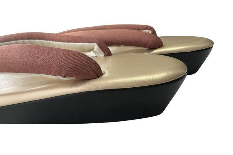May include: A pair of traditional Japanese geta sandals. The sandals feature a gold-colored platform with a black base. The thong straps are a muted reddish-brown color. The sandals are elevated and appear to be unworn.