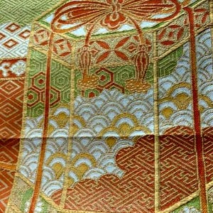 Op de afbeelding: Close-up van een gedetailleerd textiel met een herhalend patroon. Het ontwerp bevat geometrische vormen, gestileerde bloemelementen en golfpatronen in tinten groen, oranje, goud en wit. De stof heeft een getextureerde uitstraling.