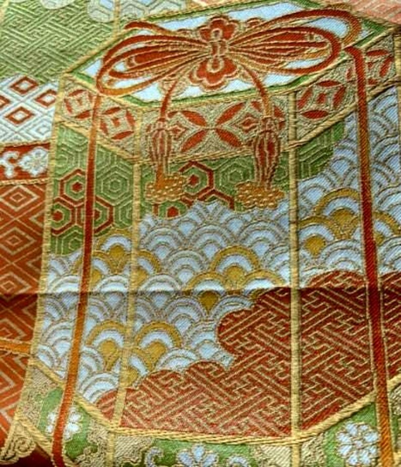 Op de afbeelding: Close-up van een gedetailleerd textiel met een herhalend patroon. Het ontwerp bevat geometrische vormen, gestileerde bloemelementen en golfpatronen in tinten groen, oranje, goud en wit. De stof heeft een getextureerde uitstraling.