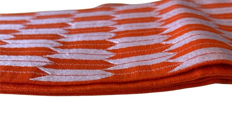May include: A vibrant orange fabric band featuring a repeating white arrow-like pattern. The material has a textured, woven appearance with a distinct horizontal orientation, showcasing a consistent geometric design across the entire surface.