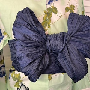 Op de afbeelding: Een donkerblauwe obi-ceintuur gestrikt in een grote, volumineuze strik met een gekreukte textuur, gedragen over een licht mintgroene kimono met een patroon van blauwe ochtendgloriebloemen en groene bladeren op een witte achtergrond.