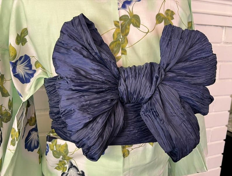 Op de afbeelding: Een donkerblauwe obi-ceintuur gestrikt in een grote, volumineuze strik met een gekreukte textuur, gedragen over een licht mintgroene kimono met een patroon van blauwe ochtendgloriebloemen en groene bladeren op een witte achtergrond.