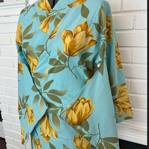 May include: A light blue kimono featuring a repeating pattern of large golden yellow flowers and muted olive green leaves. The garment has a wrap-front design with wide sleeves and a structured collar, displayed on a mannequin torso.