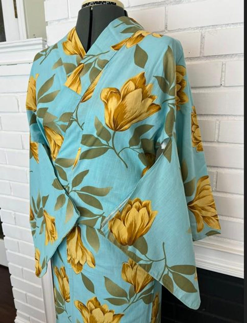 May include: A light blue kimono featuring a repeating pattern of large golden yellow flowers and muted olive green leaves. The garment has a wrap-front design with wide sleeves and a structured collar, displayed on a mannequin torso.