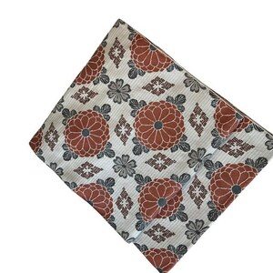 May include: A folded textile featuring a repeating pattern of large, rust-coloured chrysanthemum-like flowers with dark grey centres, interspersed with smaller grey floral motifs and geometric diamond shapes on a light, textured off-white background.