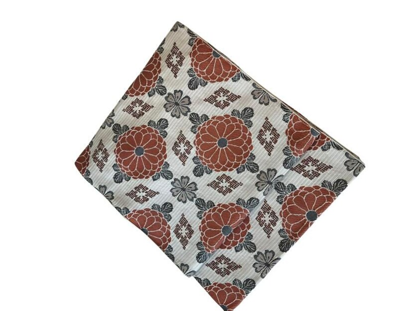 May include: A folded textile featuring a repeating pattern of large, rust-coloured chrysanthemum-like flowers with dark grey centres, interspersed with smaller grey floral motifs and geometric diamond shapes on a light, textured off-white background.
