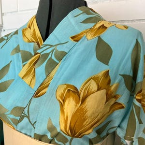 May include: A light blue kimono featuring a large-scale floral pattern in shades of golden yellow and muted olive green. The garment has a traditional wrap-style front with a V-neckline and wide sleeves, displayed on a mannequin torso.