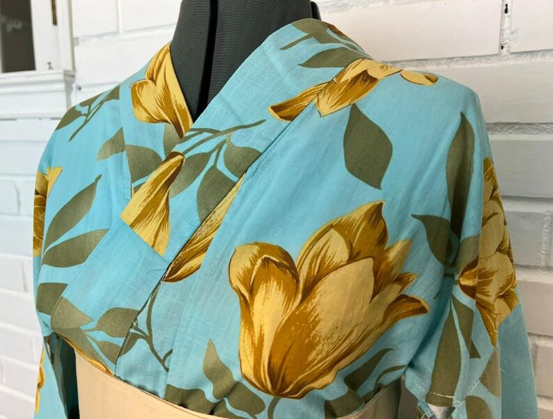 May include: A light blue kimono featuring a large-scale floral pattern in shades of golden yellow and muted olive green. The garment has a traditional wrap-style front with a V-neckline and wide sleeves, displayed on a mannequin torso.