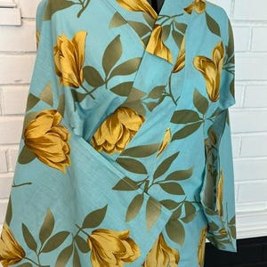 May include: A light blue kimono featuring a repeating pattern of large yellow flowers and olive green leaves. The garment has a wrap-front design with wide sleeves and a structured collar, displayed on a mannequin against a white brick background.