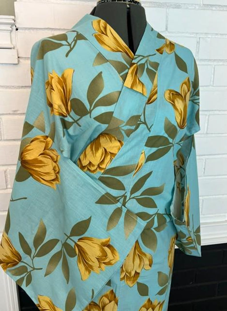 May include: A light blue kimono featuring a repeating pattern of large yellow flowers and olive green leaves. The garment has a wrap-front design with wide sleeves and a structured collar, displayed on a mannequin against a white brick background.