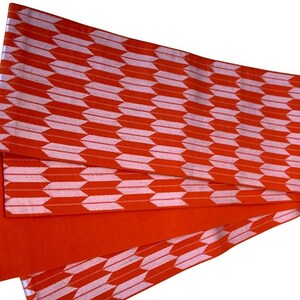May include: A rectangular fabric sash featuring a repeating pattern of light pink arrow-like shapes arranged in horizontal rows against a vibrant orange background. The textile is folded, revealing a solid orange underside and finished edges.