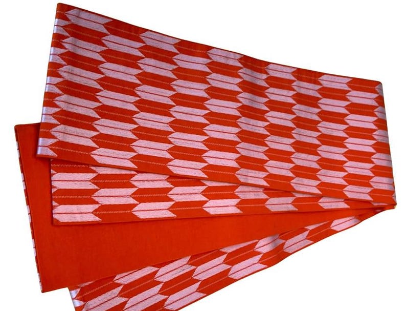 May include: A rectangular fabric sash featuring a repeating pattern of light pink arrow-like shapes arranged in horizontal rows against a vibrant orange background. The textile is folded, revealing a solid orange underside and finished edges.