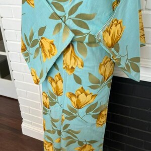 May include: A light blue kimono featuring a repeating pattern of large yellow flowers and olive green leaves. The garment has wide, structured sleeves and a wrap-front design, showcasing a vibrant floral print across the entire fabric surface.