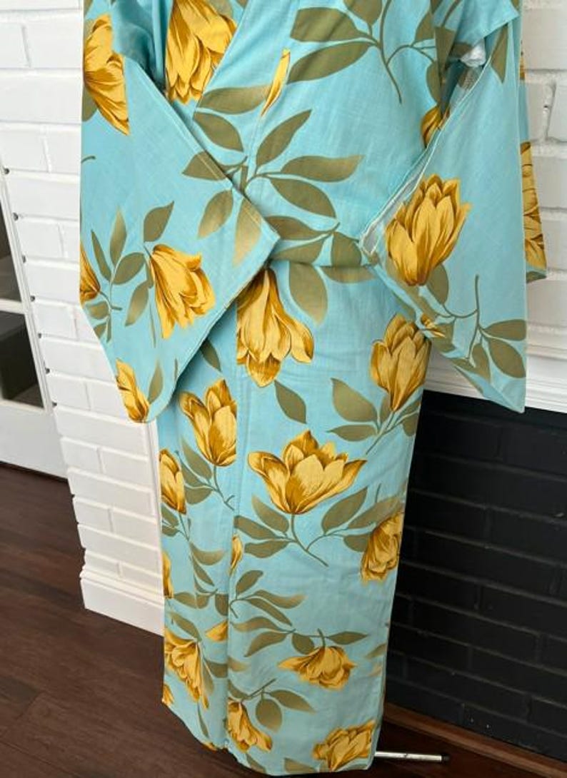 May include: A light blue kimono featuring a repeating pattern of large yellow flowers and olive green leaves. The garment has wide, structured sleeves and a wrap-front design, showcasing a vibrant floral print across the entire fabric surface.