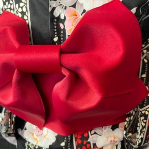 May include: A large, vibrant red fabric bow tied in a structured knot, positioned over a black garment featuring a repeating pattern of white and pink cherry blossoms and geometric lines. The bow has a smooth, matte texture and wide, folded loops.