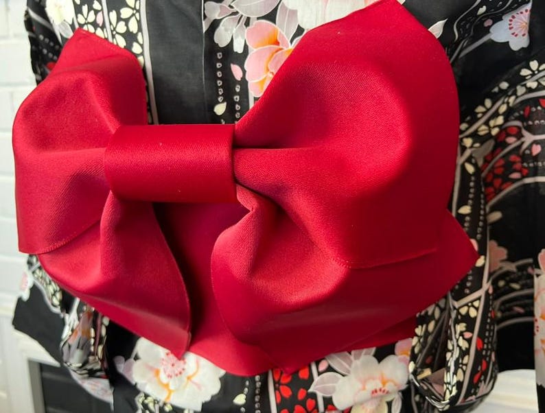 May include: A large, vibrant red fabric bow tied in a structured knot, positioned over a black garment featuring a repeating pattern of white and pink cherry blossoms and geometric lines. The bow has a smooth, matte texture and wide, folded loops.