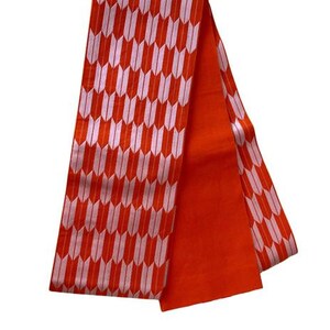 May include: A long, rectangular fabric sash featuring a vibrant orange background with a repeating pattern of light pink arrow-like shapes. One side of the sash displays this geometric design, while the other side is a solid, plain orange color.