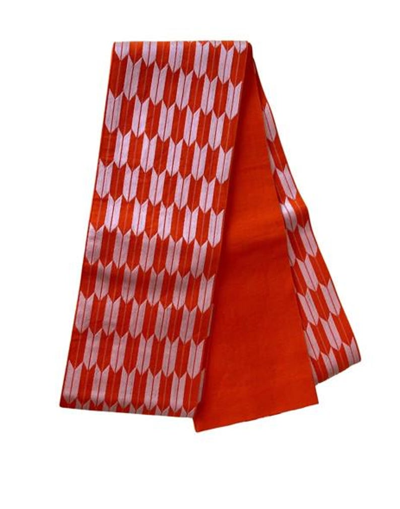 May include: A long, rectangular fabric sash featuring a vibrant orange background with a repeating pattern of light pink arrow-like shapes. One side of the sash displays this geometric design, while the other side is a solid, plain orange color.