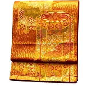 Op de afbeelding: Een gevouwen, sierlijke textielstof met een herhalend geometrisch patroon in tinten goud, oranje en groen. De stof heeft een gedetailleerd ontwerp met een centraal motief van een strik en kwasten. De algehele esthetiek suggereert een traditioneel of ceremonieel kledingstuk.
