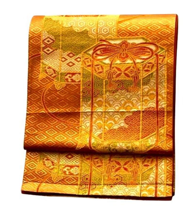 Op de afbeelding: Een gevouwen, sierlijke textielstof met een herhalend geometrisch patroon in tinten goud, oranje en groen. De stof heeft een gedetailleerd ontwerp met een centraal motief van een strik en kwasten. De algehele esthetiek suggereert een traditioneel of ceremonieel kledingstuk.