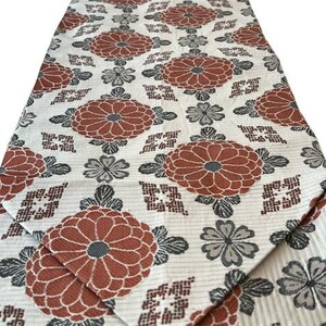 May include: A long rectangular table runner featuring a repeating pattern of large rust-orange chrysanthemum flowers and smaller grey floral accents on a cream-coloured background with a subtle horizontal ribbed texture throughout the fabric.
