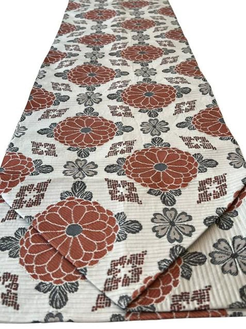May include: A long rectangular table runner featuring a repeating pattern of large rust-orange chrysanthemum flowers and smaller grey floral accents on a cream-coloured background with a subtle horizontal ribbed texture throughout the fabric.