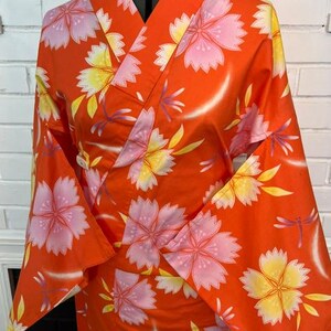 Może przedstawiać: Jaskrawopomarańczowe kimono z powtarzającym się wzorem dużych r&oacute;żowych i ż&oacute;łtych kwiat&oacute;w wiśni oraz małych fioletowych ważek. Ubi&oacute;r posiada szerokie rękawy, tradycyjny kr&oacute;j kopertowy oraz usztywniany kołnierz zaprezentowany na manekinie.