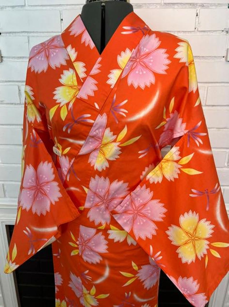 Może przedstawiać: Jaskrawopomarańczowe kimono z powtarzającym się wzorem dużych r&oacute;żowych i ż&oacute;łtych kwiat&oacute;w wiśni oraz małych fioletowych ważek. Ubi&oacute;r posiada szerokie rękawy, tradycyjny kr&oacute;j kopertowy oraz usztywniany kołnierz zaprezentowany na manekinie.