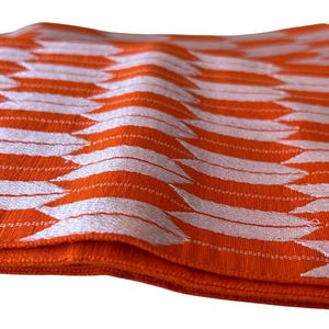 May include: A folded fabric textile featuring a vibrant orange base color with a repeating white geometric pattern of stylized arrowheads or feathers arranged in vertical columns across the textured surface of the material.