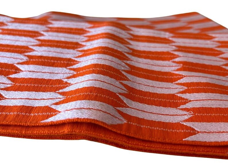 May include: A folded fabric textile featuring a vibrant orange base color with a repeating white geometric pattern of stylized arrowheads or feathers arranged in vertical columns across the textured surface of the material.