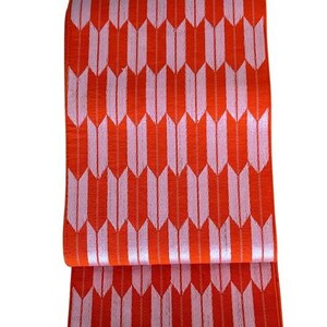 May include: A rectangular textile piece featuring a repeating pattern of vertical, stylized arrow shapes in light pink against a vibrant orange background. The fabric has a visible woven texture and is folded horizontally across the middle section.