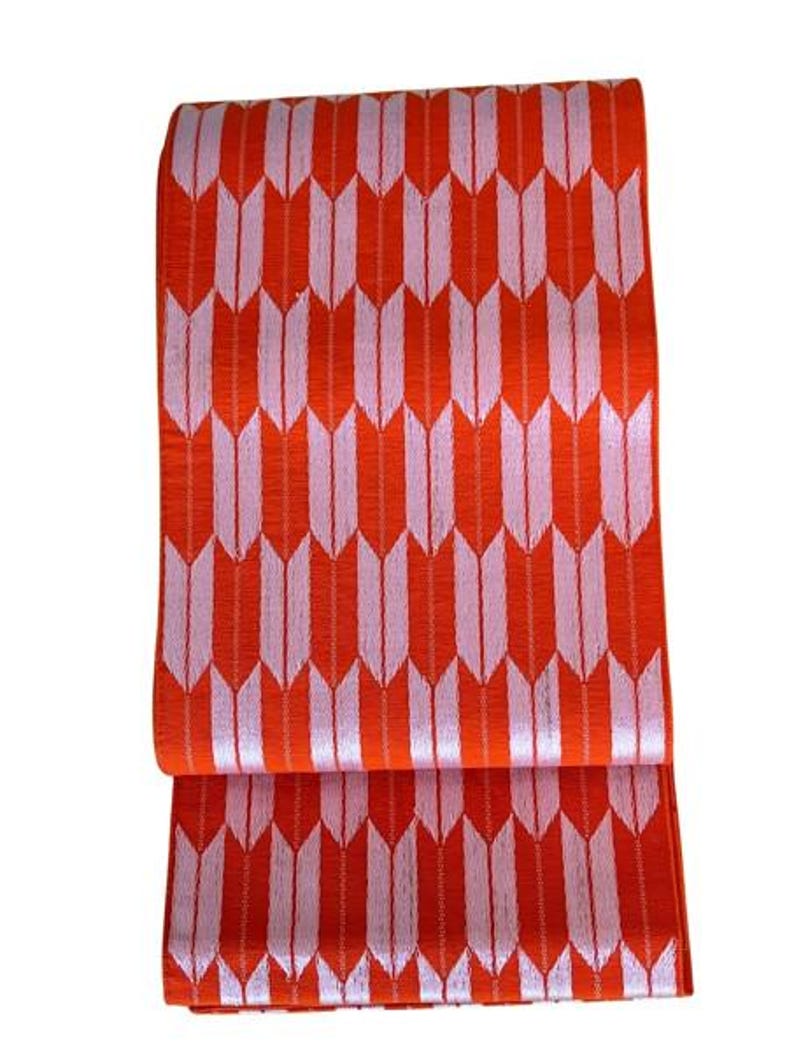 May include: A rectangular textile piece featuring a repeating pattern of vertical, stylized arrow shapes in light pink against a vibrant orange background. The fabric has a visible woven texture and is folded horizontally across the middle section.