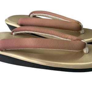 May include: A pair of traditional Japanese geta sandals. The sandals have a gold-colored base with a black sole and a pink and white fabric thong. The thong is padded for comfort. The sandals are designed to be worn outdoors.