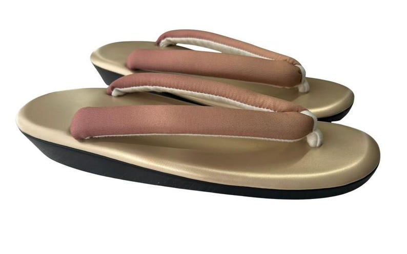 May include: A pair of traditional Japanese geta sandals. The sandals have a gold-colored base with a black sole and a pink and white fabric thong. The thong is padded for comfort. The sandals are designed to be worn outdoors.