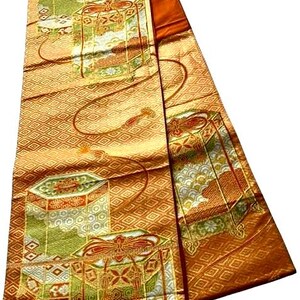 Op de afbeelding: Een gevouwen, sierlijk textiel met een herhalend geometrisch patroon in goud en oranje. De stof heeft gedetailleerde illustraties van decoratieve dozen in groen, wit en rood, met gouden accenten. Het ontwerp suggereert een traditioneel of ceremonieel thema.