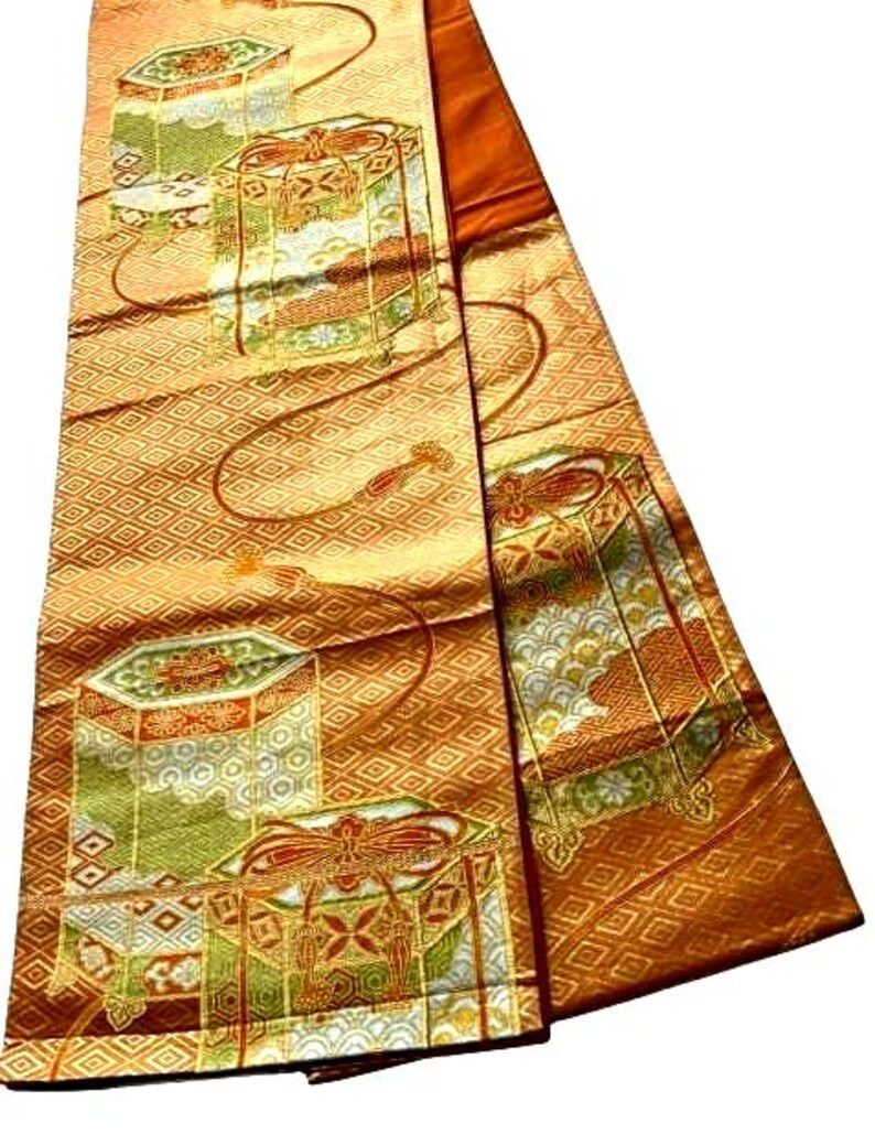 Op de afbeelding: Een gevouwen, sierlijk textiel met een herhalend geometrisch patroon in goud en oranje. De stof heeft gedetailleerde illustraties van decoratieve dozen in groen, wit en rood, met gouden accenten. Het ontwerp suggereert een traditioneel of ceremonieel thema.