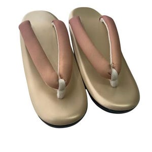 May include: A pair of traditional Japanese geta sandals. The sandals have a beige base with a black sole and a pink thong strap. The thong is attached with a white cord.