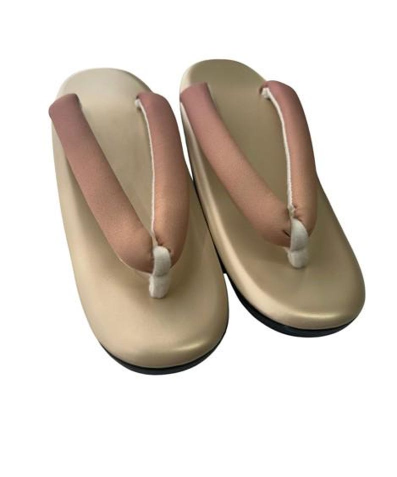 May include: A pair of traditional Japanese geta sandals. The sandals have a beige base with a black sole and a pink thong strap. The thong is attached with a white cord.
